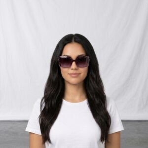 Woman with long dark hair wearing oversized purple sunglasses and a white T-shirt, standing in front of a plain backdrop.