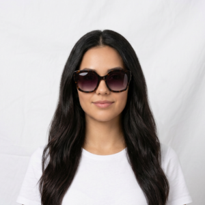 Portrait of a woman with long dark hair wearing large tortoiseshell sunglasses and a white shirt against a neutral backdrop.
